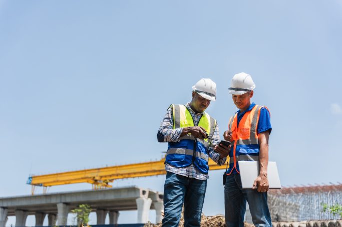 Zwei Bauarbeiter, die auf einer Baustelle zusammen auf ein Handy schauen.